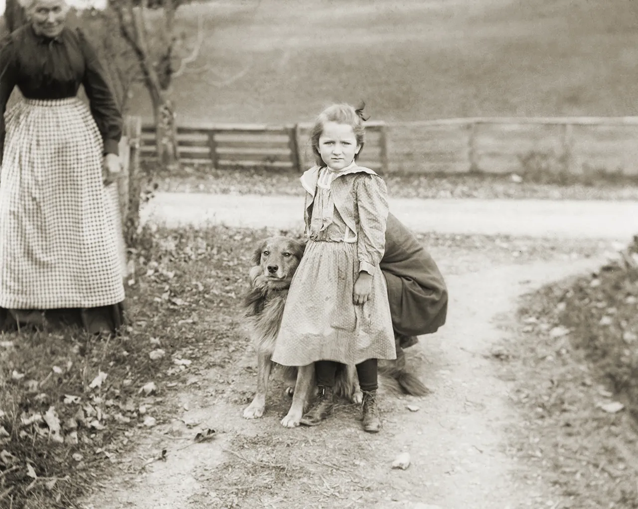 A girl stands beside a dog as a person kneels behind them.
